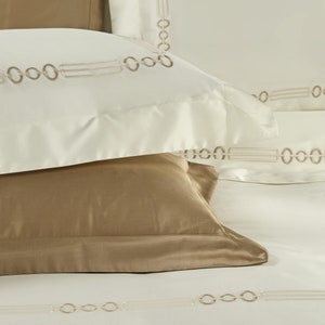 May include: A set of bedding featuring cream-colored pillowcases and a flat sheet with an embroidered pattern of interlocking circles and horizontal lines in a light brown thread. A solid tan pillowcase rests beneath the cream pillowcase.