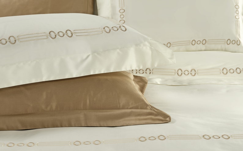 May include: A set of bedding featuring cream-colored pillowcases and a flat sheet with an embroidered pattern of interlocking circles and horizontal lines in a light brown thread. A solid tan pillowcase rests beneath the cream pillowcase.