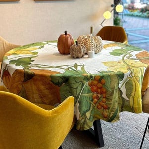 Könnte beinhalten: Ein runder Tischläufer mit einem botanischen Design mit Kürbissen, Kürbissen und Trauben in Grün-, Gelb- und Orangetönen. Dekorative Kürbisse und ein Kerzenhalter stehen auf dem Tisch. Der Tischläufer ist über einem schwarzen Tisch drapiert.