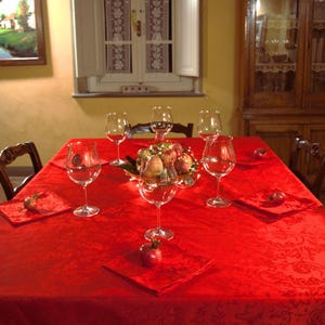 Può includere: Una tovaglia rossa vibrante con un sottile motivo damascato copre un tavolo da pranzo. Il tavolo è apparecchiato con cinque calici da vino in vetro trasparente, tovaglioli di stoffa rossa abbinati e una ciotola centrale con frutta e luci.