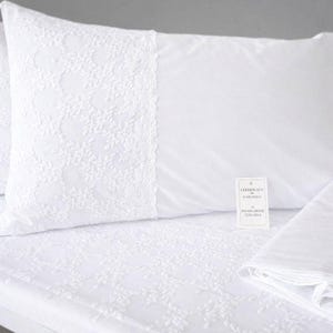 Puede incluir: Una funda de almohada y una sábana a juego de color blanco con un intrincado borde de encaje floral blanco. Una pequeña etiqueta rectangular está unida a la funda con el texto CERTIFICATO DI GARANZIA DI PRODUZIONE ITALIANA impreso en tinta negra.