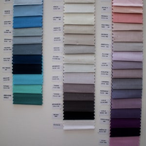 May include: A fabric swatch sample card featuring three vertical columns of rectangular textile pieces with pinked edges. The swatches display a wide spectrum of colors including blues, teals, grays, whites, pinks, purples, and blacks, each labeled with a name and code.