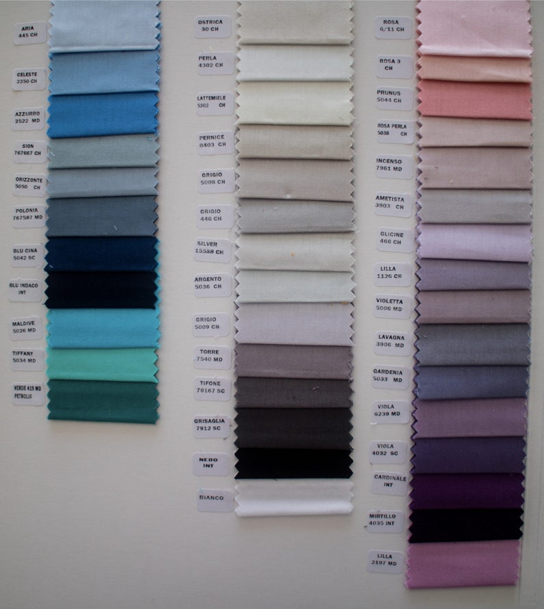 May include: A fabric swatch sample card featuring three vertical columns of rectangular textile pieces with pinked edges. The swatches display a wide spectrum of colors including blues, teals, grays, whites, pinks, purples, and blacks, each labeled with a name and code.