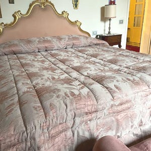 May include: A bed featuring a pink headboard with a gold ornate frame. The bed is covered with a floral patterned comforter in pink and white. The room includes a lamp, a nightstand, and decorative wall hangings. The comforter has a repeating leaf pattern.