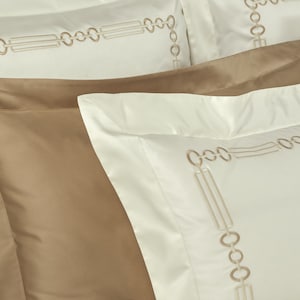 May include: A set of pillowcases featuring a smooth, solid tan pillowcase layered between two off-white pillowcases. The off-white pillowcases are decorated with a rectangular border pattern embroidered in a light tan thread, consisting of repeating circles and parallel lines.