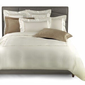 May include: A bed set featuring a cream-colored duvet cover and matching pillow shams with a delicate embroidered border of small circles and lines. The set includes solid tan accent pillowcases and rests against a dark gray upholstered headboard.