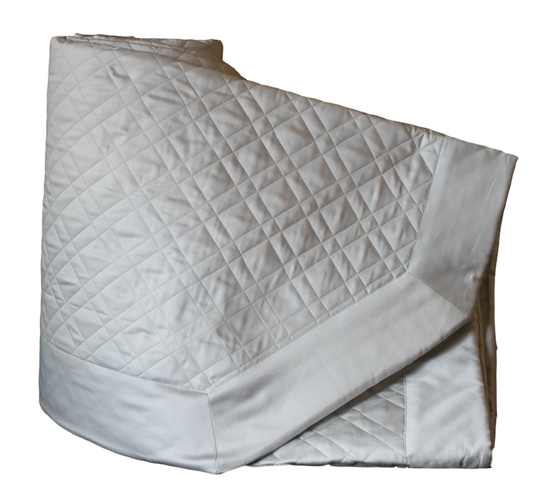 May include: A folded off-white quilt featuring a diamond-shaped quilted pattern across the center panel. The quilt has a smooth, non-quilted border along the edges, creating a clean and structured appearance for this bedding item.