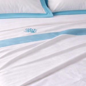 Puede incluir: Un juego de sábanas blancas con una ancha banda horizontal de color azul claro en la parte superior. La tela es lisa y nítida, acentuada por un pequeño motivo de coral bordado en azul claro centrado en la sábana bajo la banda azul.