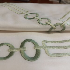May include: A white fabric textile featuring an embroidered pattern of light green circular rings and rectangular shapes. The embroidery consists of stitched lines forming a chain-like design that repeats across the smooth, folded surface of the material.