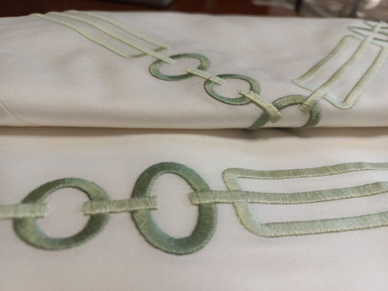 May include: A white fabric textile featuring an embroidered pattern of light green circular rings and rectangular shapes. The embroidery consists of stitched lines forming a chain-like design that repeats across the smooth, folded surface of the material.