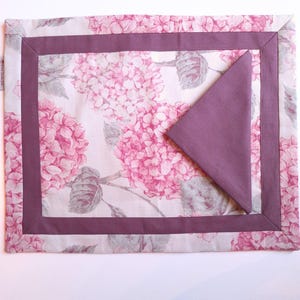 Peut inclure: Un set de table rectangulaire orné d'un motif floral d'hortensias roses et blancs avec une bordure unie mauve foncé. Une serviette triangulaire assortie de couleur mauve foncé est pliée et posée sur le set de table pour un ensemble coordonné.