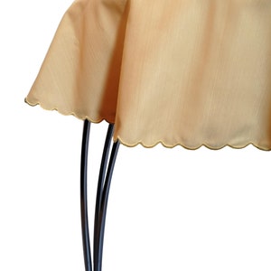 May include: A solid light gold tablecloth with a subtle vertical texture and a scalloped edge. The fabric drapes over a dark, curved metal stand, showcasing the smooth, consistent color and the decorative, rounded hemline of the textile.
