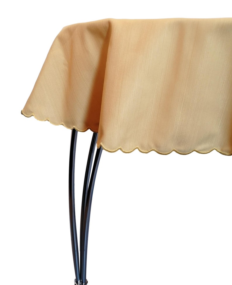 May include: A solid light gold tablecloth with a subtle vertical texture and a scalloped edge. The fabric drapes over a dark, curved metal stand, showcasing the smooth, consistent color and the decorative, rounded hemline of the textile.
