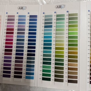 May include: A color chart for Aurifil thread featuring multiple columns of thread samples arranged by hue. Each sample is a small rectangular swatch of colored thread labeled with a corresponding numerical code on a white paper background.
