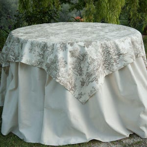May include: A round table with a cream-coloured tablecloth and a decorative overlay. The overlay has a detailed toile pattern in brown and cream, showing scenes of people and nature. The table is set outdoors.