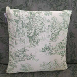 Puede incluir: Un coj&iacute;n cuadrado con base de tela blanca y un patr&oacute;n toile de jouy verde. El dise&ntilde;o muestra varias figuras en escenas al aire libre, incluyendo personas bailando, sentadas a mesas y &aacute;rboles, con una peque&ntilde;a etiqueta en la esquina superior izquierda.