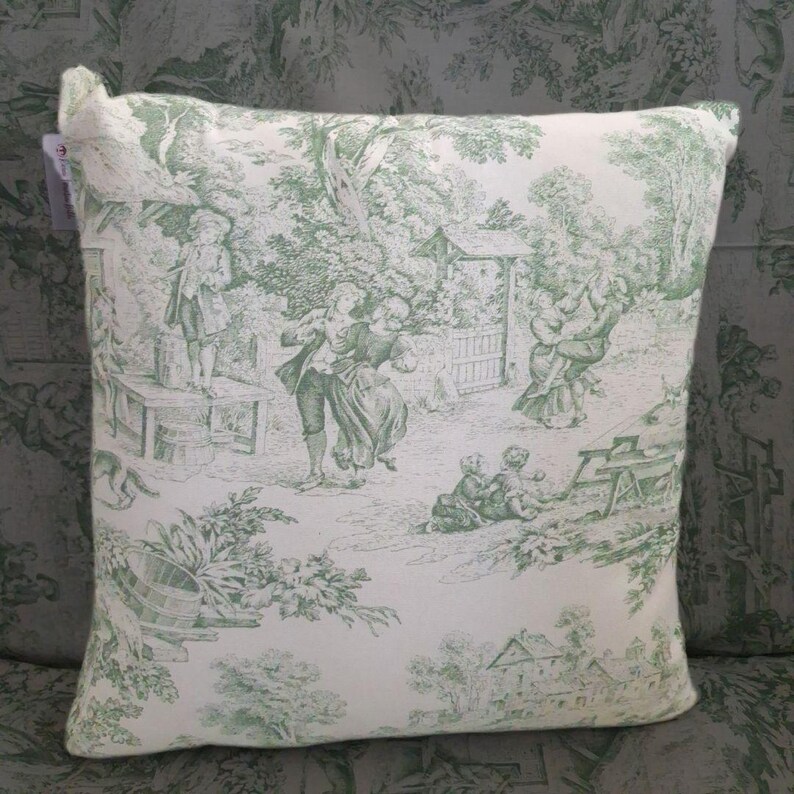 Puede incluir: Un coj&iacute;n cuadrado con base de tela blanca y un patr&oacute;n toile de jouy verde. El dise&ntilde;o muestra varias figuras en escenas al aire libre, incluyendo personas bailando, sentadas a mesas y &aacute;rboles, con una peque&ntilde;a etiqueta en la esquina superior izquierda.