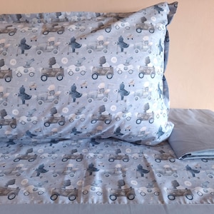 May include: Light blue bedding set featuring a repeating pattern of bears driving vintage cars, clouds, and buttons. The pillowcase is filled and has a folded edge. The flat sheet is folded and visible in the lower right corner.