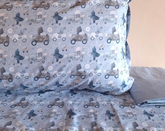 May include: A light grey pillowcase and matching bed sheet set featuring a repeating pattern of illustrated bears driving vintage-style cars and riding scooters, interspersed with small circular motifs on a soft grey fabric background.