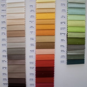 May include: A fabric swatch sample card featuring three vertical columns of rectangular textile strips in a wide spectrum of colors. Each swatch is labeled with a specific color name and code printed on small white rectangular paper tags.
