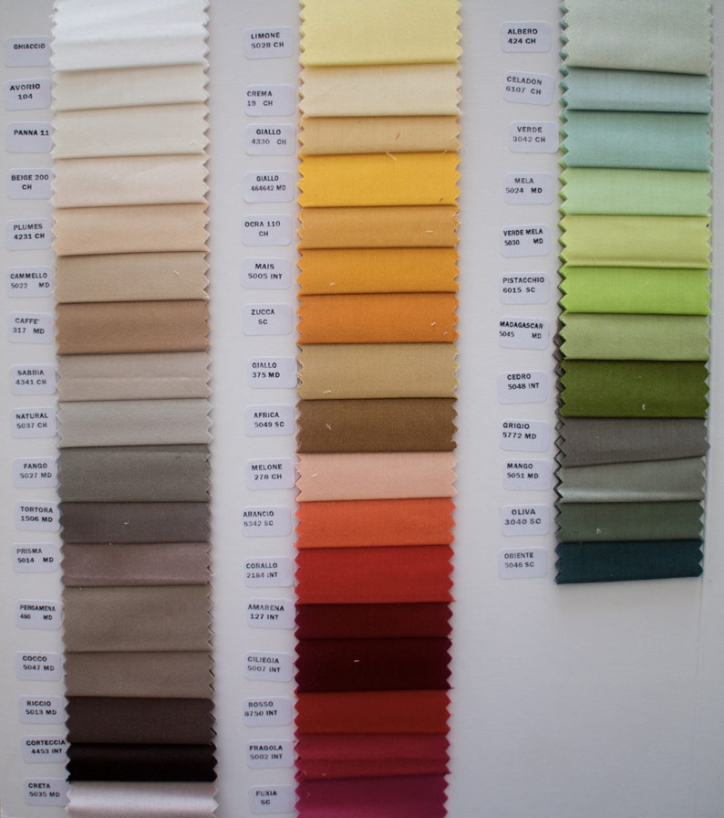 May include: A fabric swatch sample card featuring three vertical columns of rectangular textile strips in a wide spectrum of colors. Each swatch is labeled with a specific color name and code printed on small white rectangular paper tags.