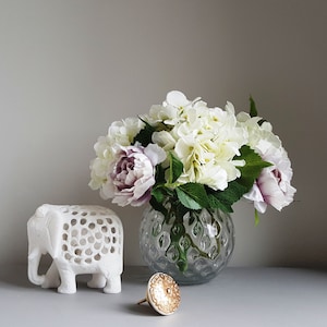 May include: A white ceramic elephant figurine with a decorative cutout design sits on a table next to a clear glass vase filled with white and purple flowers. A gold decorative knob is also on the table.