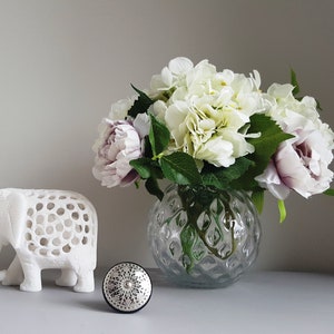 May include: A white ceramic elephant figurine with a decorative cut-out design sits on a table next to a clear glass vase with a textured surface. The vase holds a bouquet of white and purple flowers.