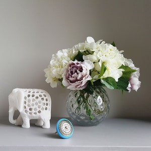 May include: A white ceramic elephant figurine with intricate cutouts sits on a grey surface next to a blue and gold decorative knob. A clear glass vase with a textured surface holds a bouquet of white and purple flowers.