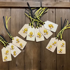 Puede incluir: Un conjunto de diez etiquetas de regalo de madera con diseños de abejas amarillas y negras. Cada etiqueta tiene una cinta negra y amarilla.