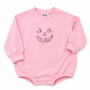 May include: A light pink long-sleeve romper featuring a crew neckline and ribbed cuffs. The center of the chest displays an embroidered jack-o'-lantern face with triangular eyes and a wide grin, filled with a colorful floral pattern on a white background.