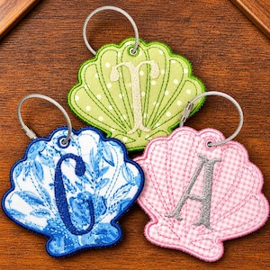 May include: Three seashell-shaped keychains with embroidered letters. One is light green with white polka dots and a beige "T". Another is blue and white floral with a blue "C". The third is pink gingham with a silver "A". Each has a silver wire loop.