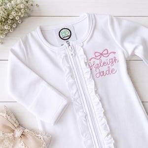 May include: A white long-sleeve zippered sleeper featuring a vertical ruffle detail along the front closure. The garment is embroidered with a pink bow and the name Haleigh Jade. A tag inside the neck indicates a size of 3M.