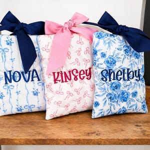 May include: Three rectangular fabric pouches featuring embroidered names. The left pouch is white with blue ribbons and the name NOVA. The center pouch is white with pink bows and the name KINSEY. The right pouch has blue floral patterns and the name SHELBY.
