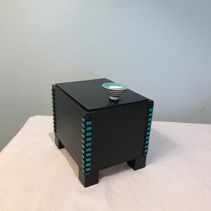 May include: A black wooden box with a lid, featuring turquoise accents along the edges. The box has a small, round handle on top with a blue and white striped design. The box is elevated on four short legs.