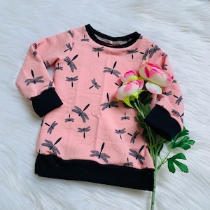 Può includere: Felpa rosa chiaro con polsini e orlo neri, decorata con libellule grigie e linee tratteggiate. Il colletto è nero. L'indumento è esposto su una superficie bianca e soffice.