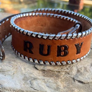 May include: A brown leather bracelet with the name "RUBY" embossed in black lettering. The edges are trimmed with silver-colored metal and white stitching. The bracelet is displayed on a textured stone surface.