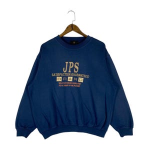 May include: A navy blue sweatshirt with the text "JPS SATISFACTION GUARANTEED BRAND" in a gold color. The sweatshirt has a crew neck and long sleeves. The text "THE ACTIVE PERSONS CASUAL WEAR FOR A CHASER OF PURE PLEASURE" is also visible.
