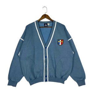 May include: A light blue cardigan sweater with a V-neck and white trim. The sweater has a button-up front and a patch on the chest with a logo. The sleeves are long, and the sweater is hanging on a wooden hanger.