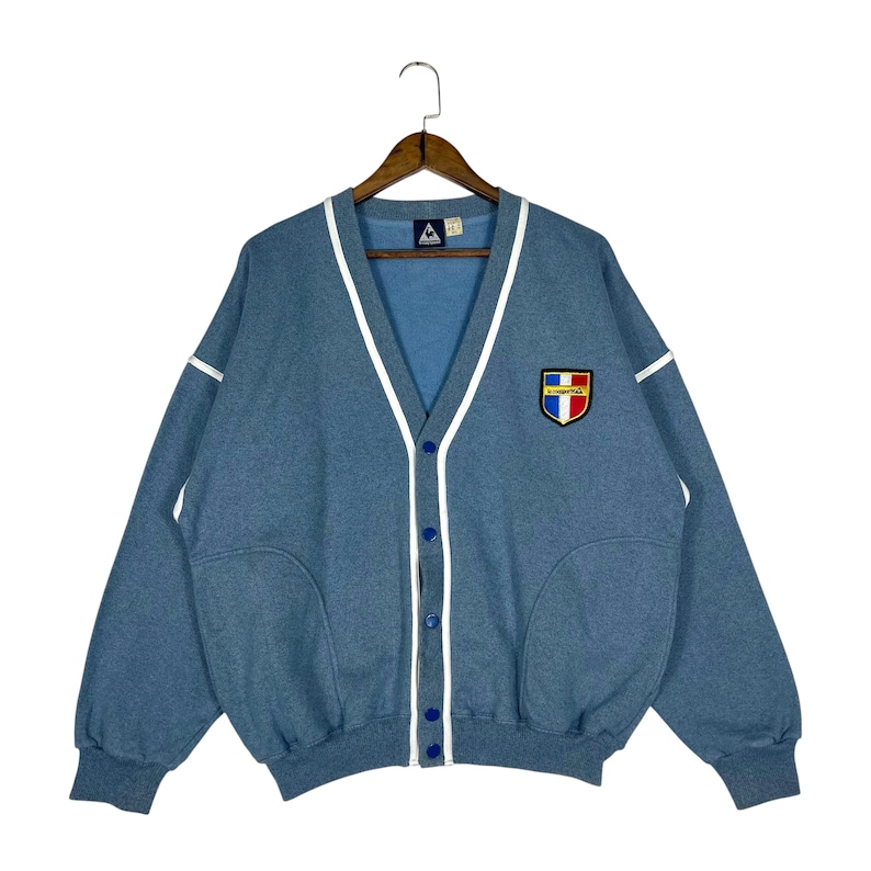 May include: A light blue cardigan sweater with a V-neck and white trim. The sweater has a button-up front and a patch on the chest with a logo. The sleeves are long, and the sweater is hanging on a wooden hanger.