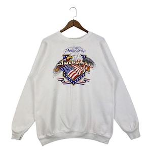 Puede incluir: Sudadera blanca con un estampado gráfico de un águila sosteniendo una bandera estadounidense. El gráfico también incluye el texto "Proud to be American".