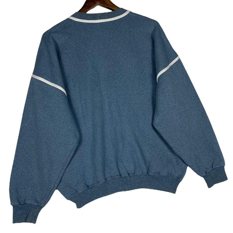 May include: A blue sweatshirt with white trim on the collar and sleeves. The sweatshirt has a crew neck and long sleeves. The cuffs and hem are a darker shade of blue. The garment is hanging on a wooden hanger.