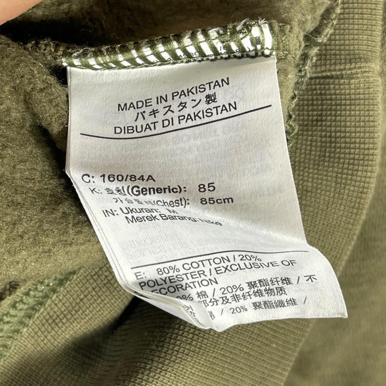 Op de afbeelding: Detailopname van een label op een groen kledingstuk. Het label toont "MADE IN PAKISTAN" in het Engels, Japans en Indonesisch. Het bevat maatdetails, waaronder een borstomvang van 85 cm, en de stofsamenstelling: 80% katoen, 20% polyester.
