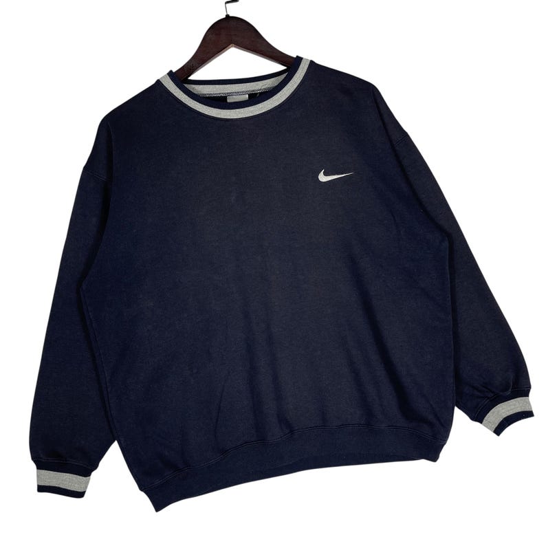 May include: Navy blue Nike sweatshirt with a white embroidered swoosh logo on the chest. The crew neck and cuffs have a gray and white striped trim. The sweatshirt is hanging on a wooden hanger.