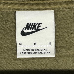 Op de afbeelding: Close-up van een wit label met het zwarte Nike-logo en de tekst "NIKE". Onder het logo staan drie maatlabels "M". Het label luidt ook "MADE IN PAKISTAN" en "FABRIQUE AU PAKISTAN". De achtergrond is olijfgroen.