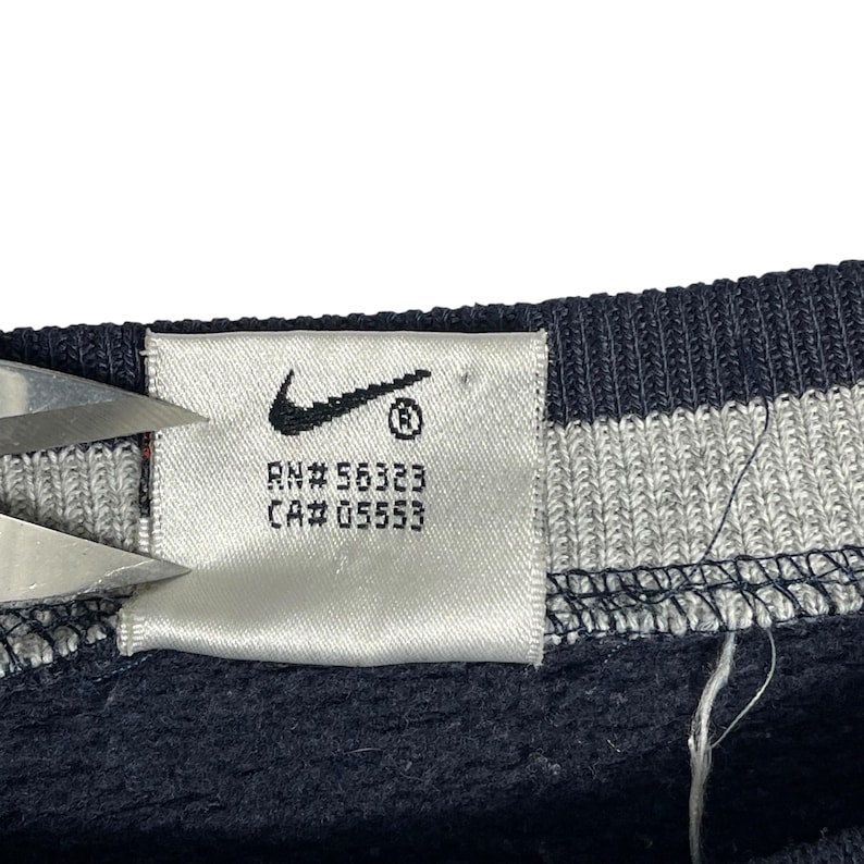 May include: Close-up of a white tag with the Nike swoosh logo and the text "RN# 56323 CA# 05553". The tag is attached to a navy blue garment with white and gray stripes.