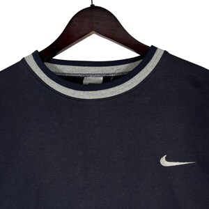 May include: Navy blue crew neck sweatshirt with a gray and white striped collar. A small white Nike swoosh logo is on the left chest. The sweatshirt is hanging on a wooden hanger.