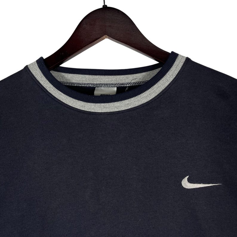 May include: Navy blue crew neck sweatshirt with a gray and white striped collar. A small white Nike swoosh logo is on the left chest. The sweatshirt is hanging on a wooden hanger.