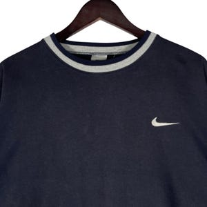 May include: Navy blue crew neck sweatshirt with a gray and white striped collar. A small white Nike swoosh logo is embroidered on the chest. The sweatshirt is hanging on a wooden hanger.