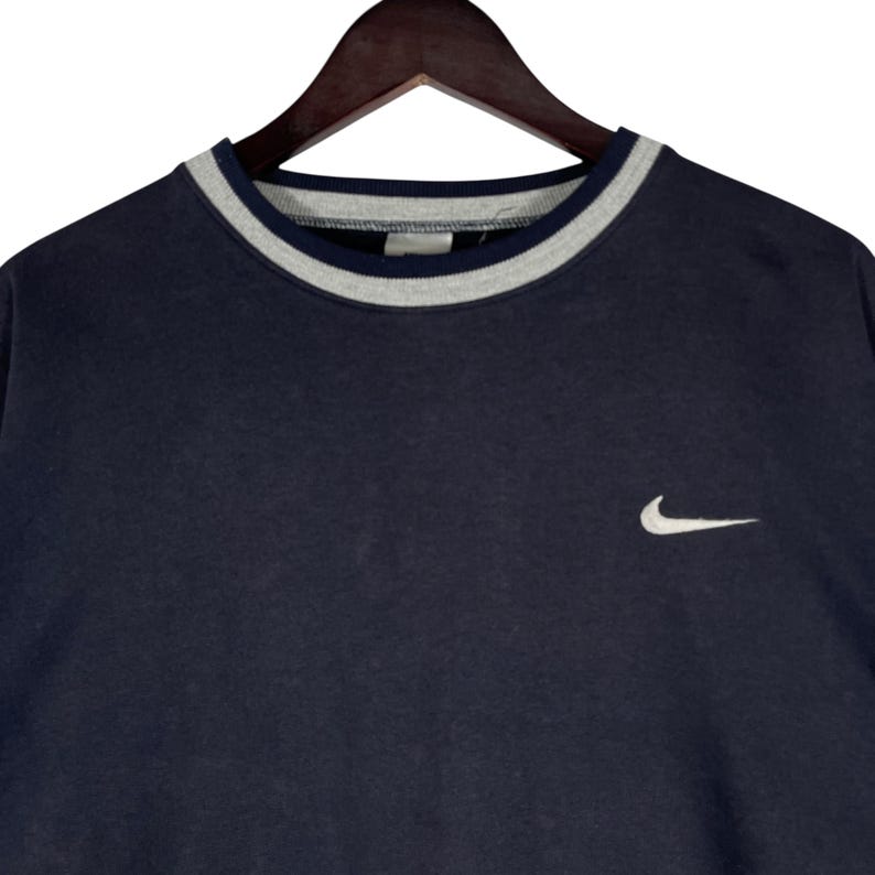 May include: Navy blue crew neck sweatshirt with a gray and white striped collar. A small white Nike swoosh logo is embroidered on the chest. The sweatshirt is hanging on a wooden hanger.