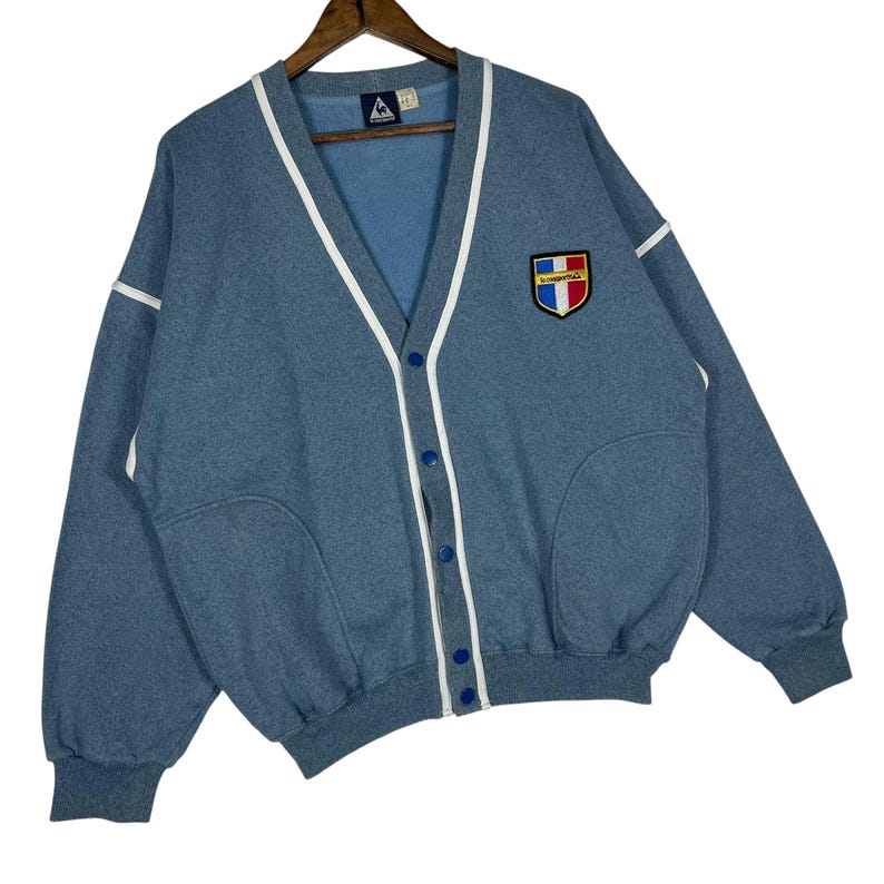 May include: A light blue cardigan sweater with white trim and a button-up front. The sweater has a patch on the chest with a shield design in red, white, blue, and yellow. The sweater is hanging on a wooden hanger.